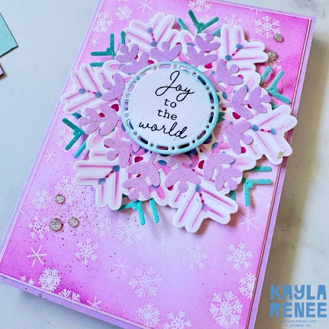Angled view of the handmade snowflake card showing dimension in the layered die cuts and shimmer accents. The circular sentiment ‘Joy to the world’ is framed by white stitching detail against a pink snowy background.