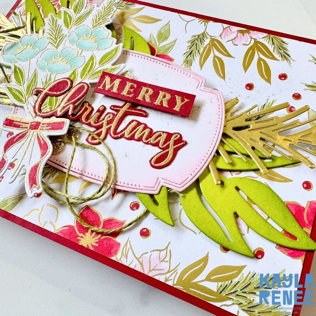 Close-up view of the Christmas card front showing intricate gold detailing, green foliage, and layered die cuts. The ‘Merry Christmas’ sentiment is framed on a pink label with a red and gold bow and twine accent.
