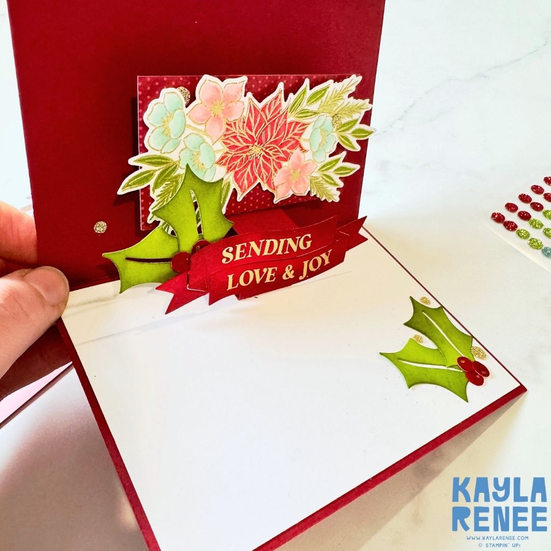 Inside view of the pop-up Christmas card featuring a red background and a dimensional floral arrangement with pastel poinsettias and blue flowers. The sentiment ‘Sending Love & Joy’ appears on a red banner with green holly leaves.