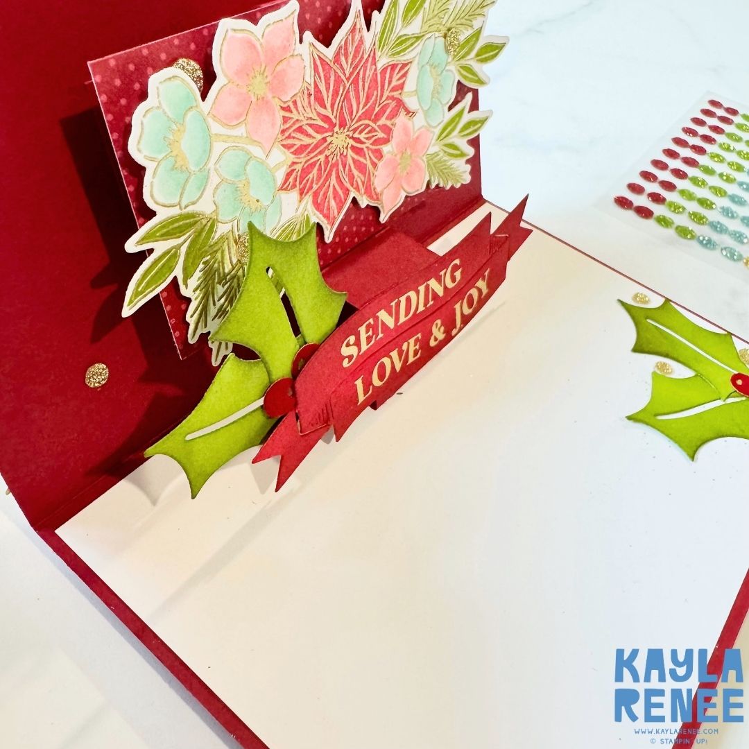 Angle view of the inside pop-up card showing layered flowers and holly leaves. The red and gold banner sentiment ‘Sending Love & Joy’ stands out at the center with festive green accents.