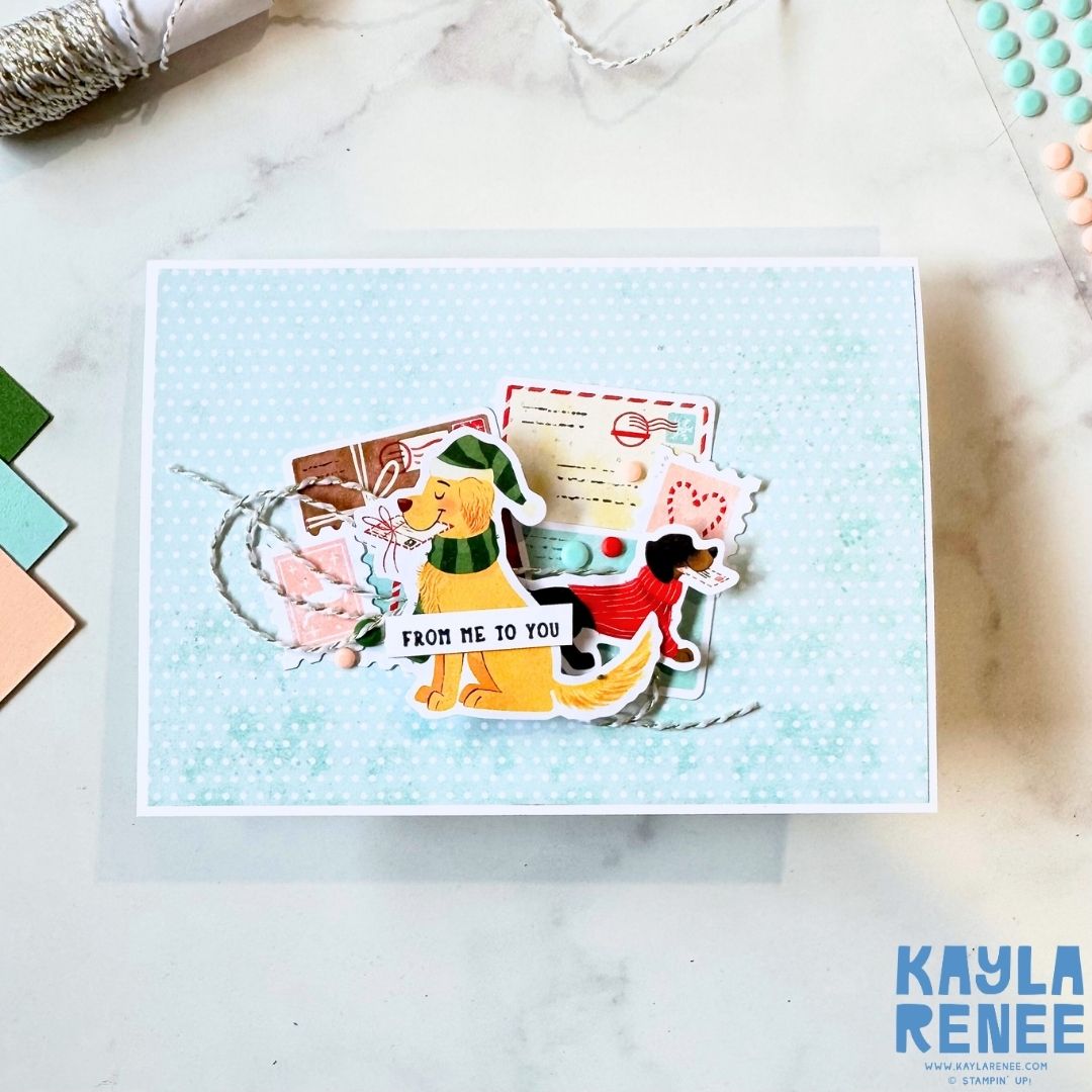 Handmade Christmas card featuring two illustrated dogs wearing festive scarves and hats, surrounded by postage-style die cuts, pastel envelopes, and twine. Sentiment reads ‘From me to you’ on a light blue polka dot background.