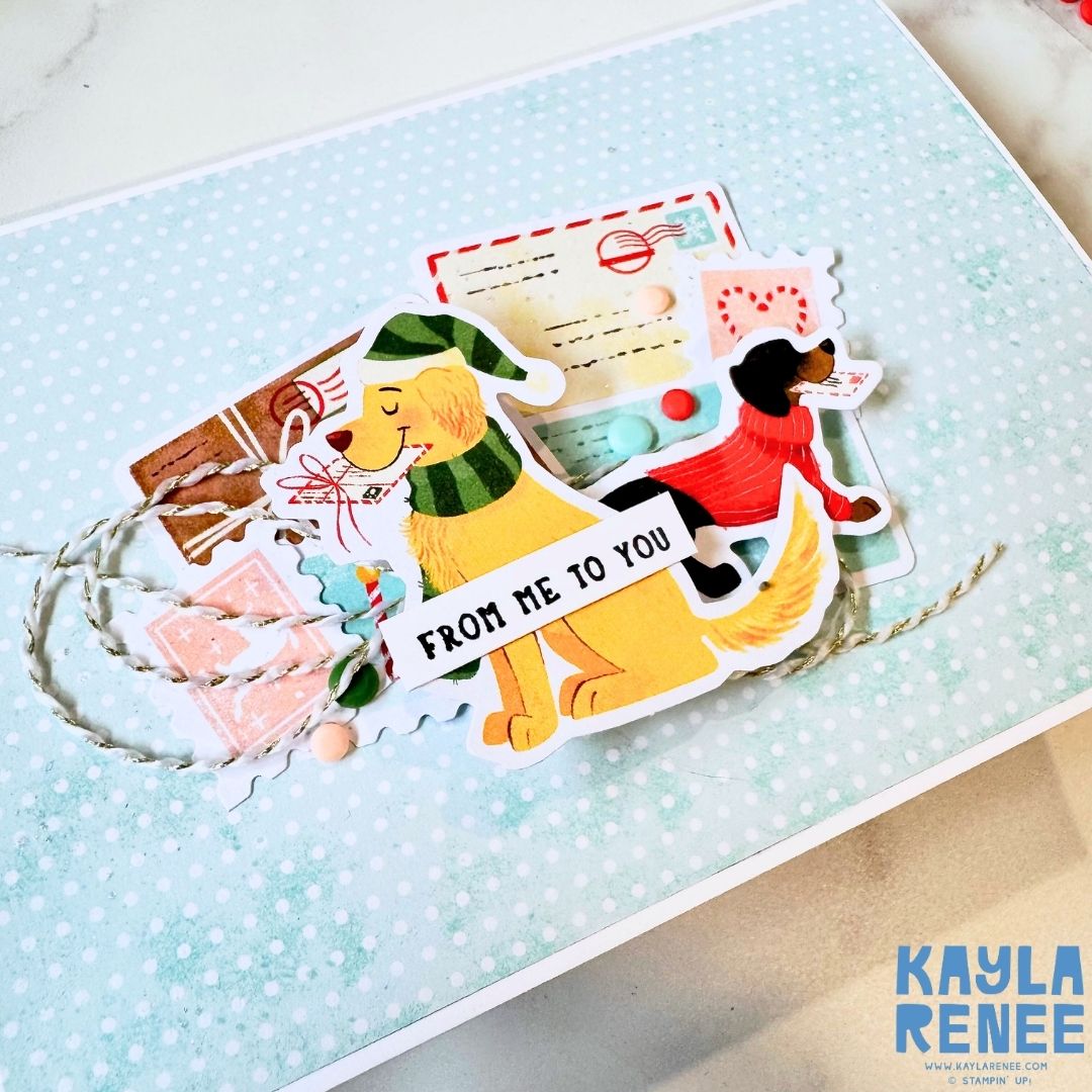 Close-up of a handmade Christmas card with layered die cuts of dogs delivering mail, pastel embellishments, and the sentiment ‘From me to you.’ The background features soft blue polka dots.