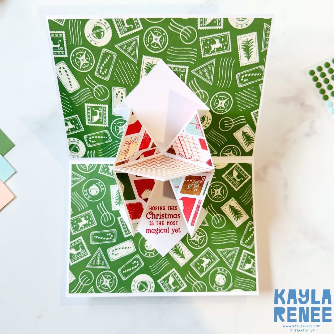 Inside view of a pop-up Christmas card with green patterned paper featuring white postage designs. The center pop-up cube displays red and white holiday patterns with the sentiment ‘Hoping this Christmas is the most magical yet.’