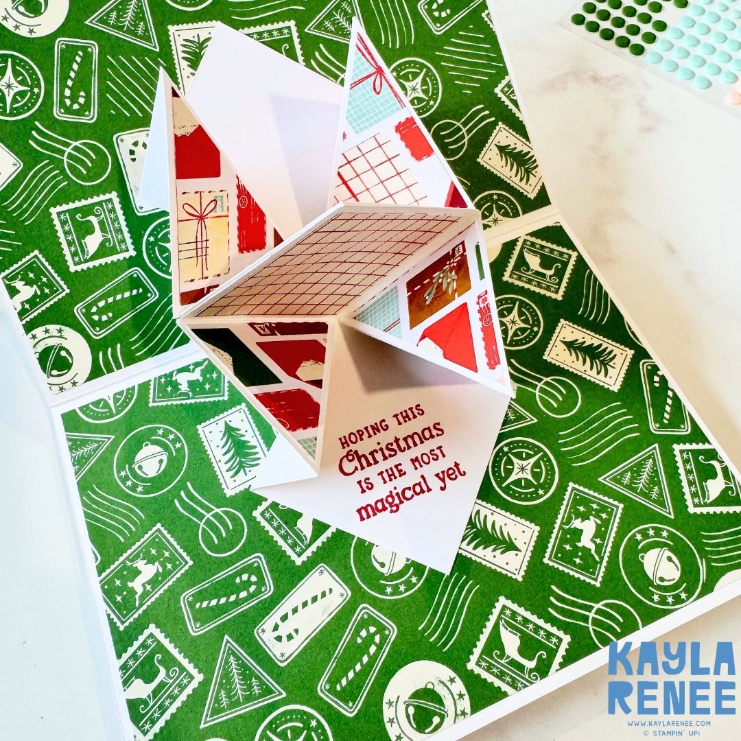 Angled view of the pop-up card showing a 3D cube design decorated with Christmas-themed paper and the red sentiment ‘Hoping this Christmas is the most magical yet.’ Green patterned paper with festive icons fills the background.