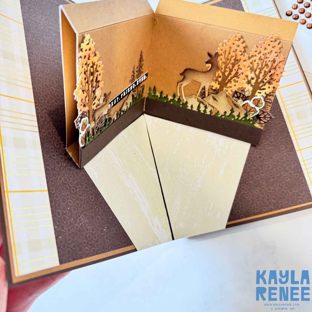 Close-up of the 3D pop-up scene featuring die-cut trees in orange and yellow tones, a deer, and forest elements like acorns and leaves, arranged against a warm brown background.