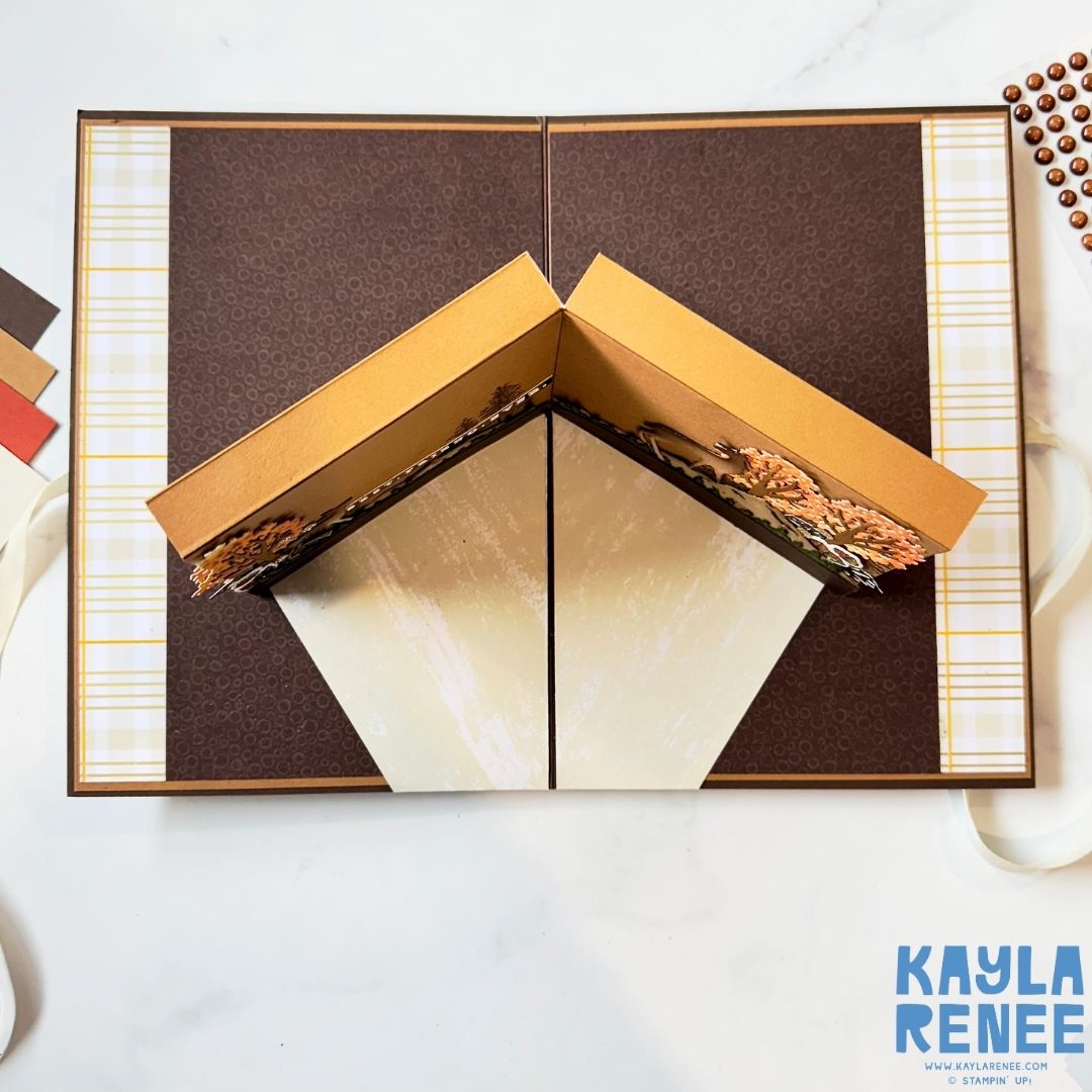 Top-down view of the open card showing the folded 3D pop-up mechanism forming a V-shape with woodland elements on each side. The card is layered with brown patterned and plaid papers.