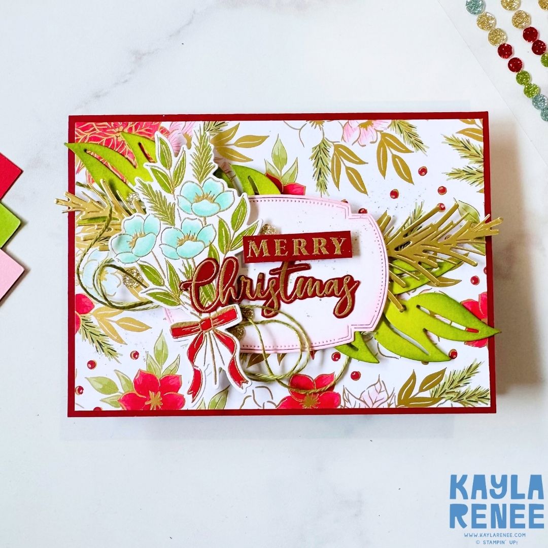 Handmade Christmas card with a red base and a festive floral background in gold, green, and red. The front design features layered die cuts of blue flowers, green pine branches, and gold accents surrounding a sentiment that reads ‘Merry Christmas’ in red and gold.