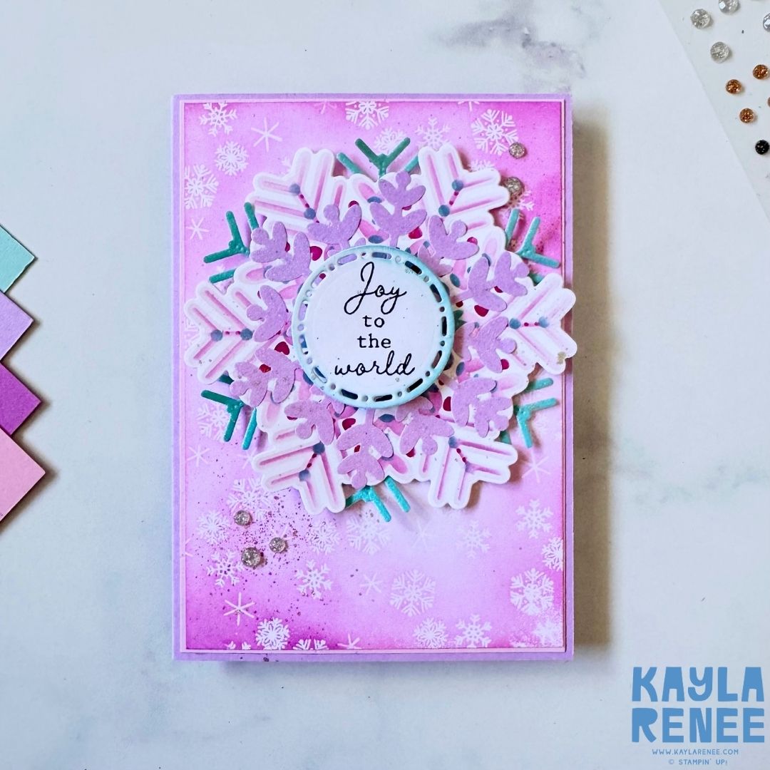 Handmade Christmas card featuring a large layered snowflake die cut in soft pink, white, and teal tones on a pink ink-blended background with stamped snowflakes. The center sentiment reads ‘Joy to the world.’ Accented with sequins and glitter dots.