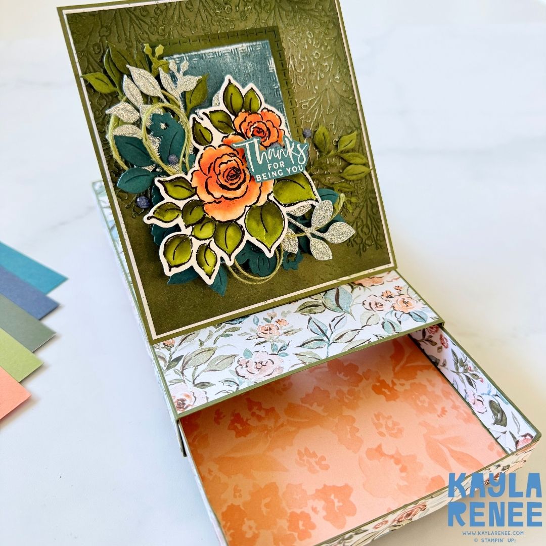 Handmade pop-up drawer card featuring floral patterned paper with a raised card front. The top panel has an olive green embossed background with layered die-cut leaves and orange watercolor roses. The sentiment reads ‘Thanks for being you.’ The drawer below is lined with peach patterned paper.