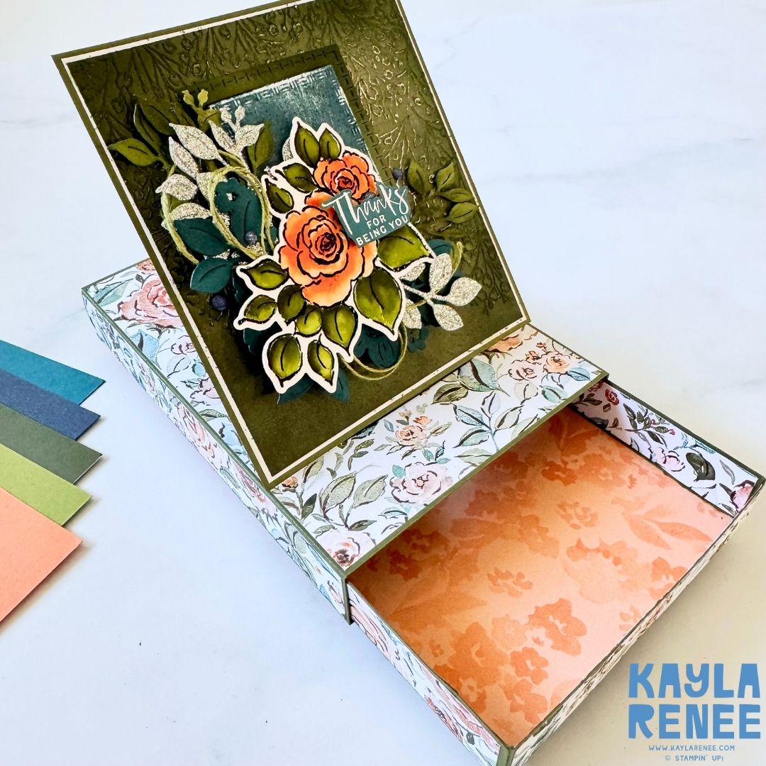 Angle view of a handmade pop-up drawer card showing the top floral arrangement of orange roses and green leaves with the sentiment ‘Thanks for being you.’ The drawer compartment is pulled open, revealing a peach floral interior.