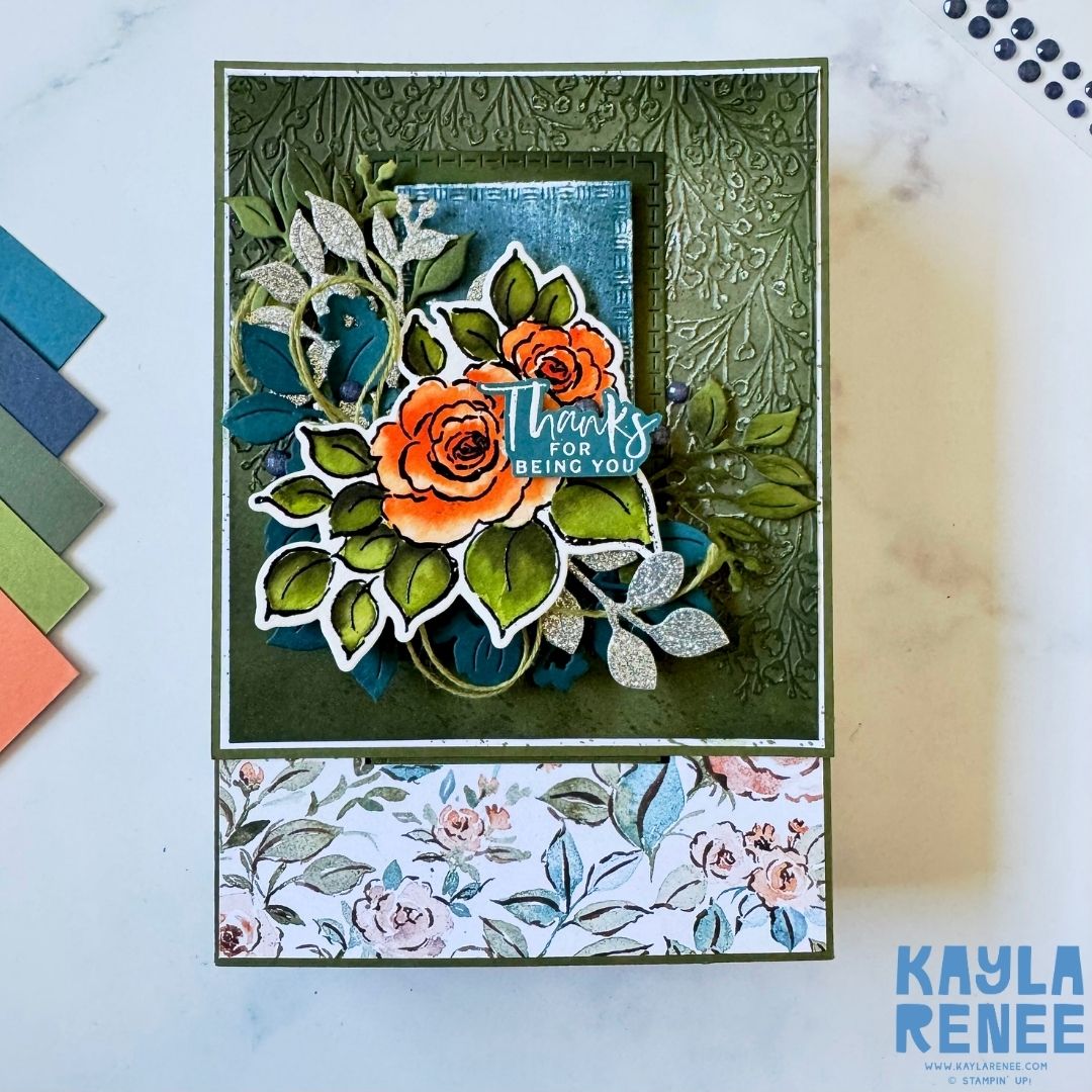 Top view of the handmade pop-up drawer card with intricate floral detailing on an embossed olive background. The central design includes orange roses, green and glittery silver leaves, and a blue sentiment label that reads ‘Thanks for being you.’ A floral patterned border decorates the bottom edge.