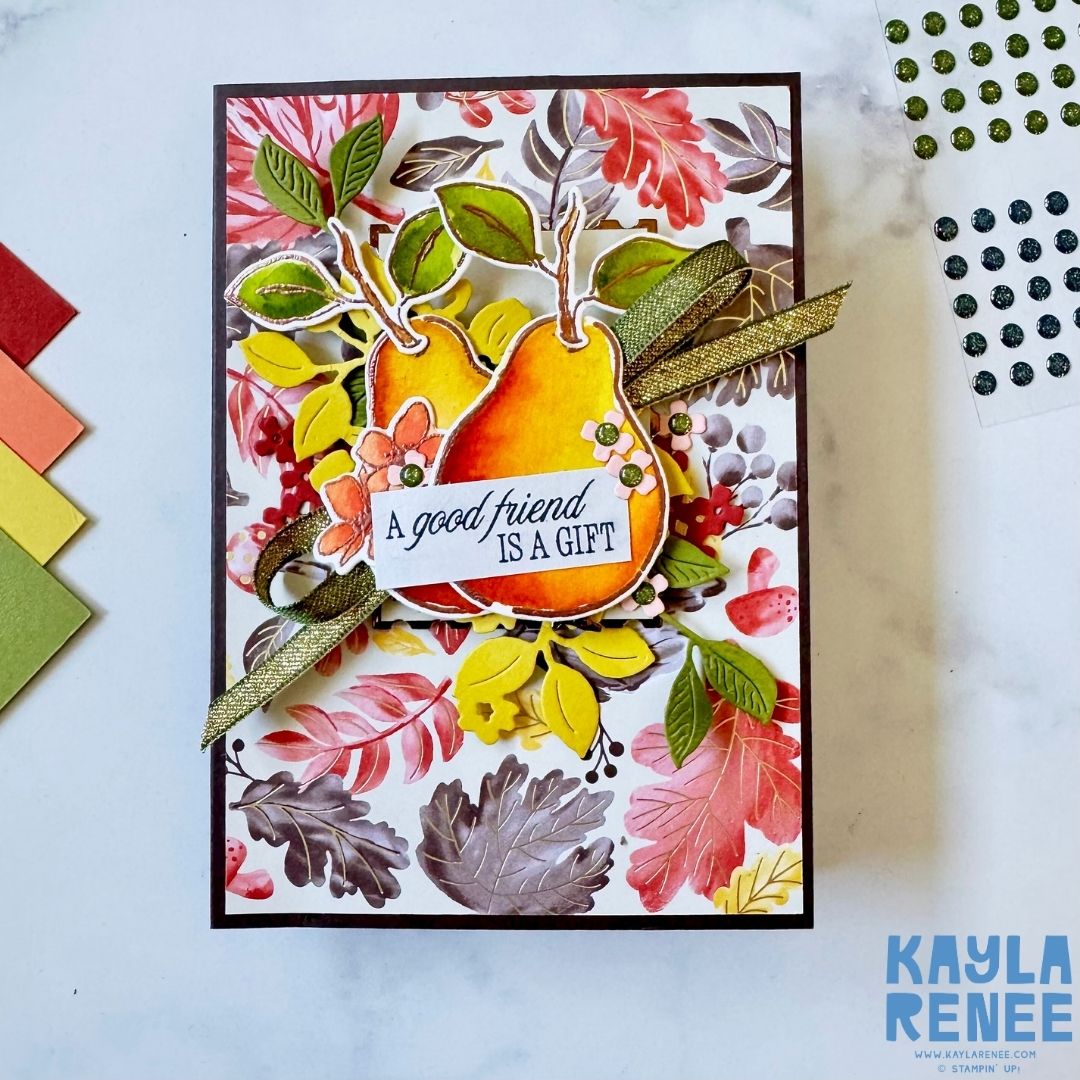 Handmade friendship card featuring two watercolour pears in yellow and orange tones with layered green leaves and ribbon detail. The sentiment reads ‘A good friend is a gift,’ placed over a vibrant leafy background in autumn shades of red, yellow, and brown.