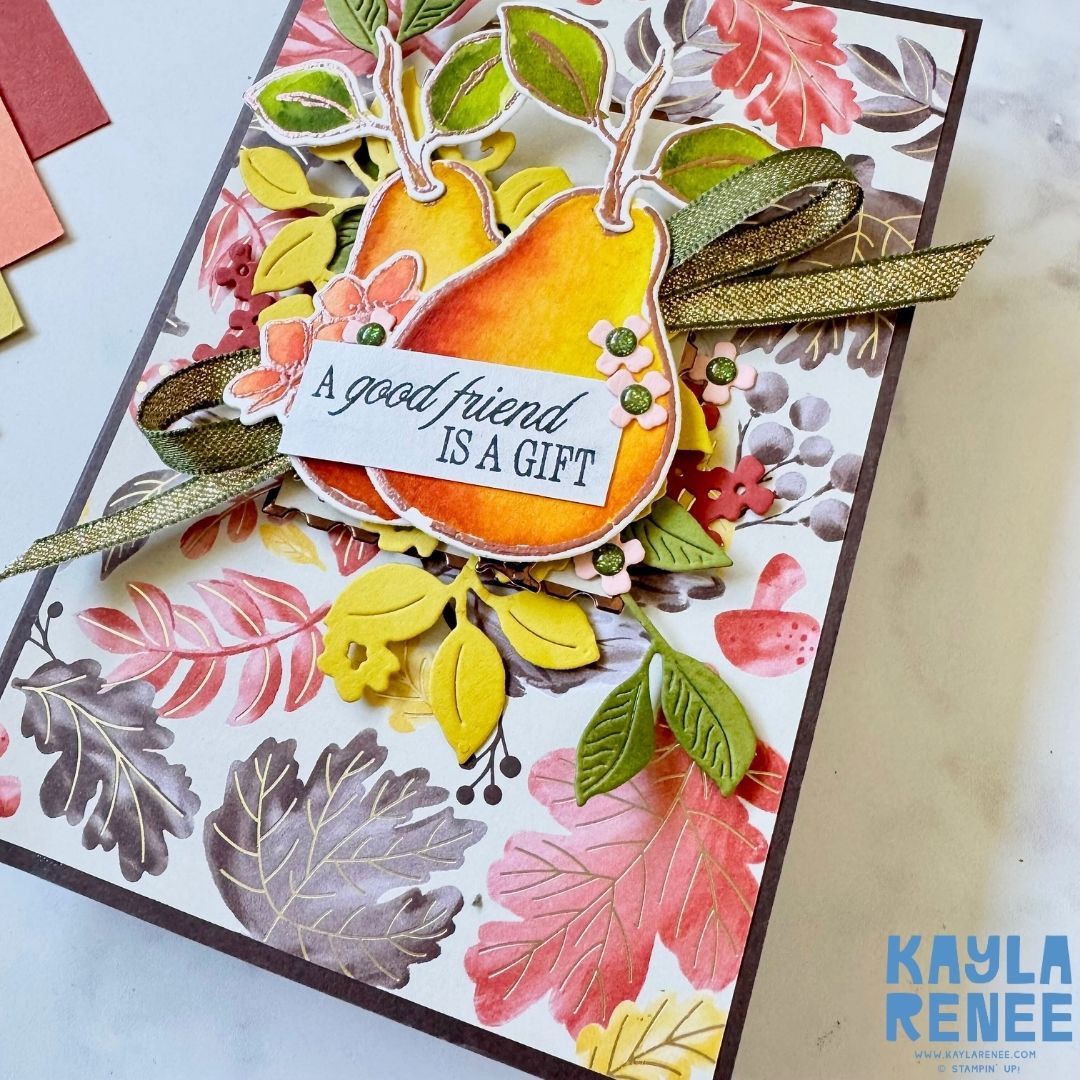 Angle view of the handmade pear-themed card showing dimensional layers of die-cut leaves, orange and yellow pears, and a metallic green ribbon. The sentiment ‘A good friend is a gift’ sits at the centre.