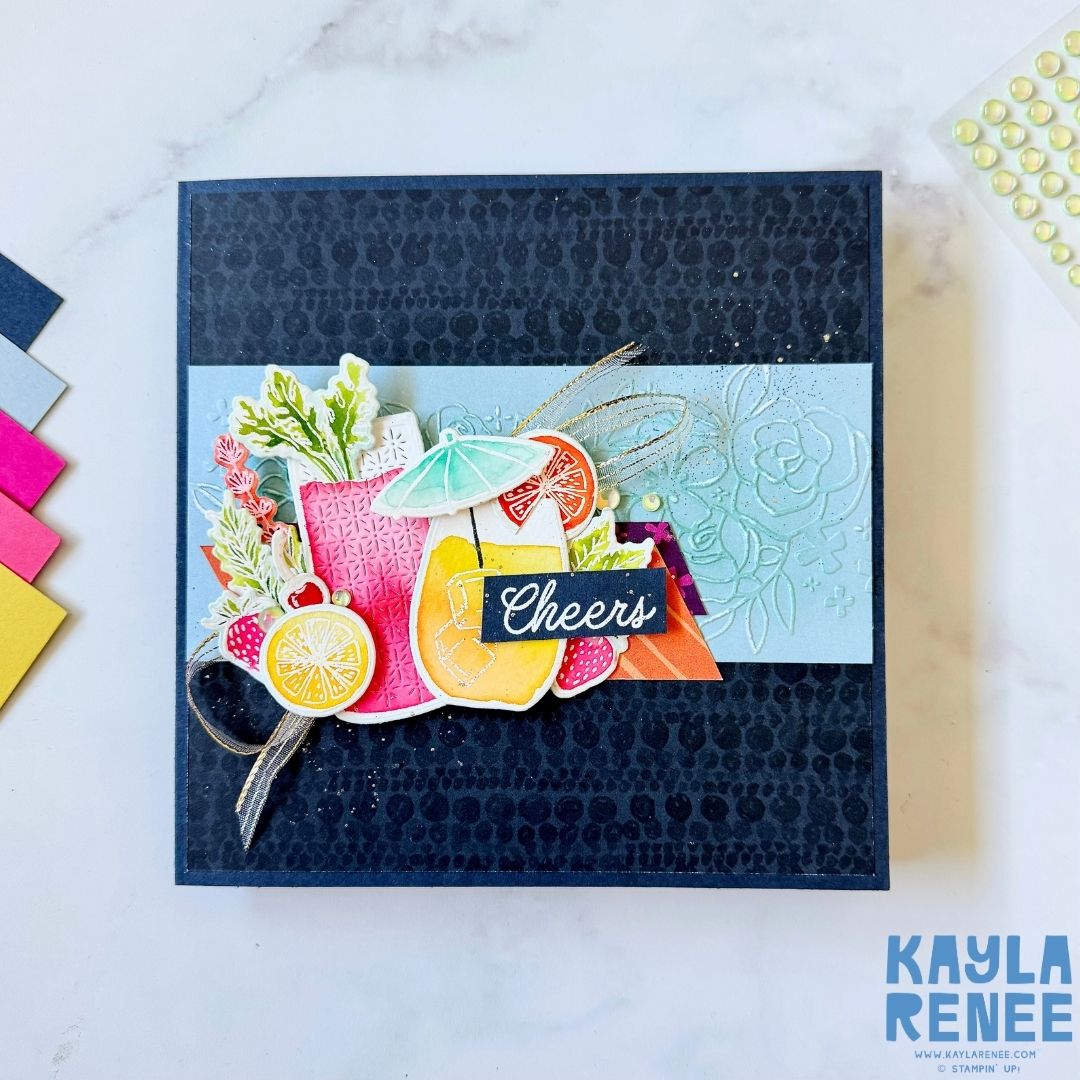 Handmade card with a navy blue embossed background and a colorful arrangement of tropical cocktail die cuts featuring oranges, lemons, and drinks under a paper umbrella. The sentiment ‘Cheers’ is displayed in white on a navy banner across the center.
