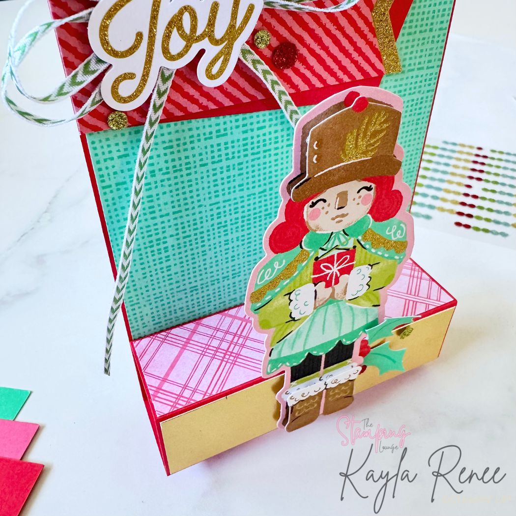 Angled close-up of a handmade step card with a whimsical nutcracker figure dressed in teal and lime green, accented with gold foil and glitter. Red and green patterned papers and a ‘Joy’ sentiment embellish the top panel.
