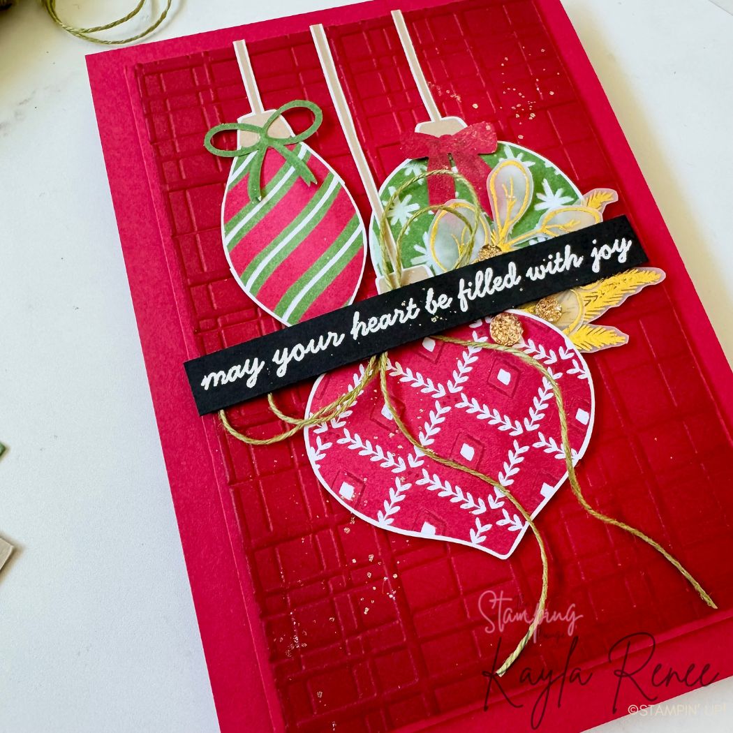 Close-up of the same handmade Christmas card, highlighting the three patterned ornaments, gold bow accents, embossed red background, and gold twine loops. The layered textures and embossing are clearly visible, along with gold splatter details.