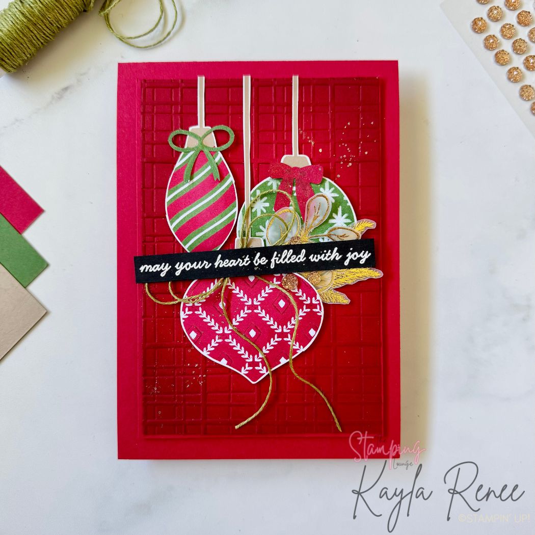 A handmade Christmas card featuring three hanging ornaments in festive greens, reds, and gold. The ornaments are layered over a textured deep red embossed background. A black sentiment strip across the center reads “may your heart be filled with joy” in white script. Gold twine accents and glittery splashes add shimmer. Card is displayed on a white background with threads and cardstock swatches nearby.