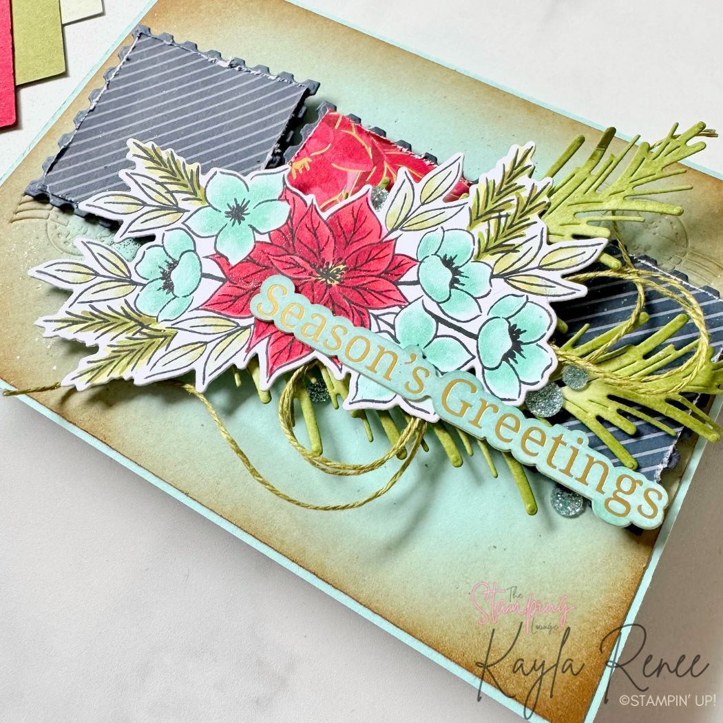 Close-up view of the same handmade Christmas card, highlighting the dimensional floral arrangement of poinsettias, aqua blossoms, and pine sprigs. The background includes stitched postage-style squares in navy and red patterned designs. Gold twine and die-cut pine elements peek from behind the florals, with the “Season's Greetings” sentiment layered on top.