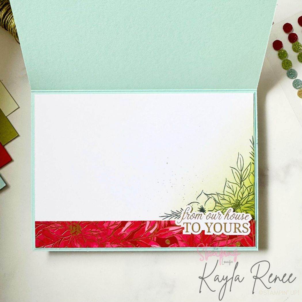 Inside panel of the handmade Christmas card featuring a white insert bordered with light aqua cardstock. The bottom edge has a red poinsettia patterned paper strip and light ink blending with stamped foliage on the lower right corner. The sentiment reads, “From our house to yours.” The design is clean and festive with soft colouring and botanical accents.