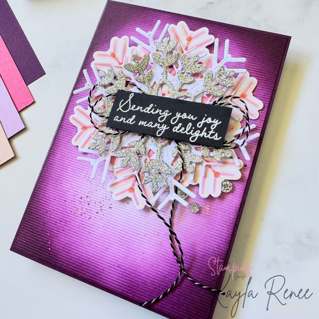 A handmade card featuring a dramatic deep-purple ink-blended background with subtle lined texture. The centre showcases a large layered snowflake built from pink and white die-cut pieces and glittery silver accents. A black sentiment panel reads “Sending you joy and many delights” in white script, wrapped with black-and-white twine. Small glitter dots add sparkle. Soft purple cardstock and coordinating cardstock swatches sit to the left.