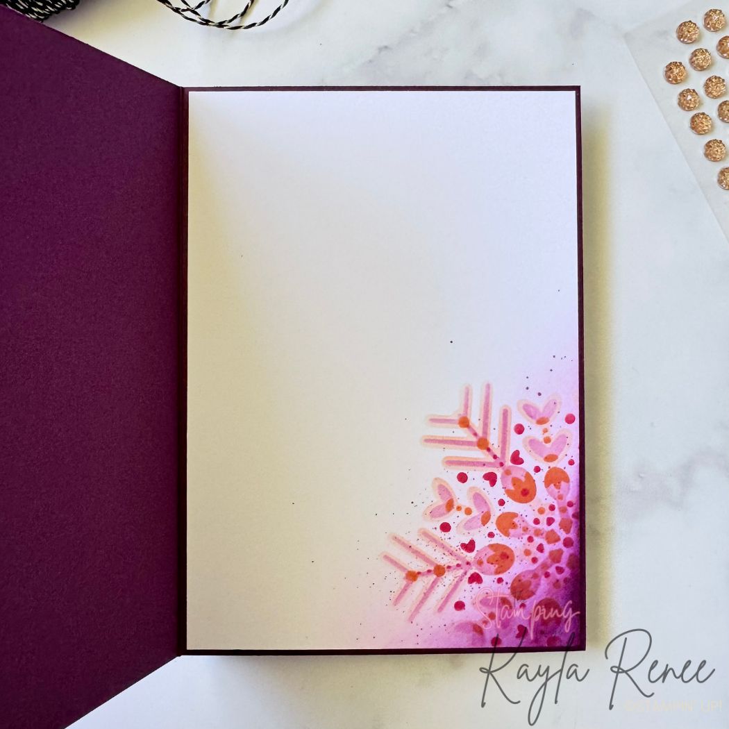 Inside of the handmade card showing a clean white insert. The bottom-right corner features bright pink and coral stencilled snowflake details with splattered ink accents, blending softly into the white space. The card base is deep purple and embellishment sheets can be seen to the side.