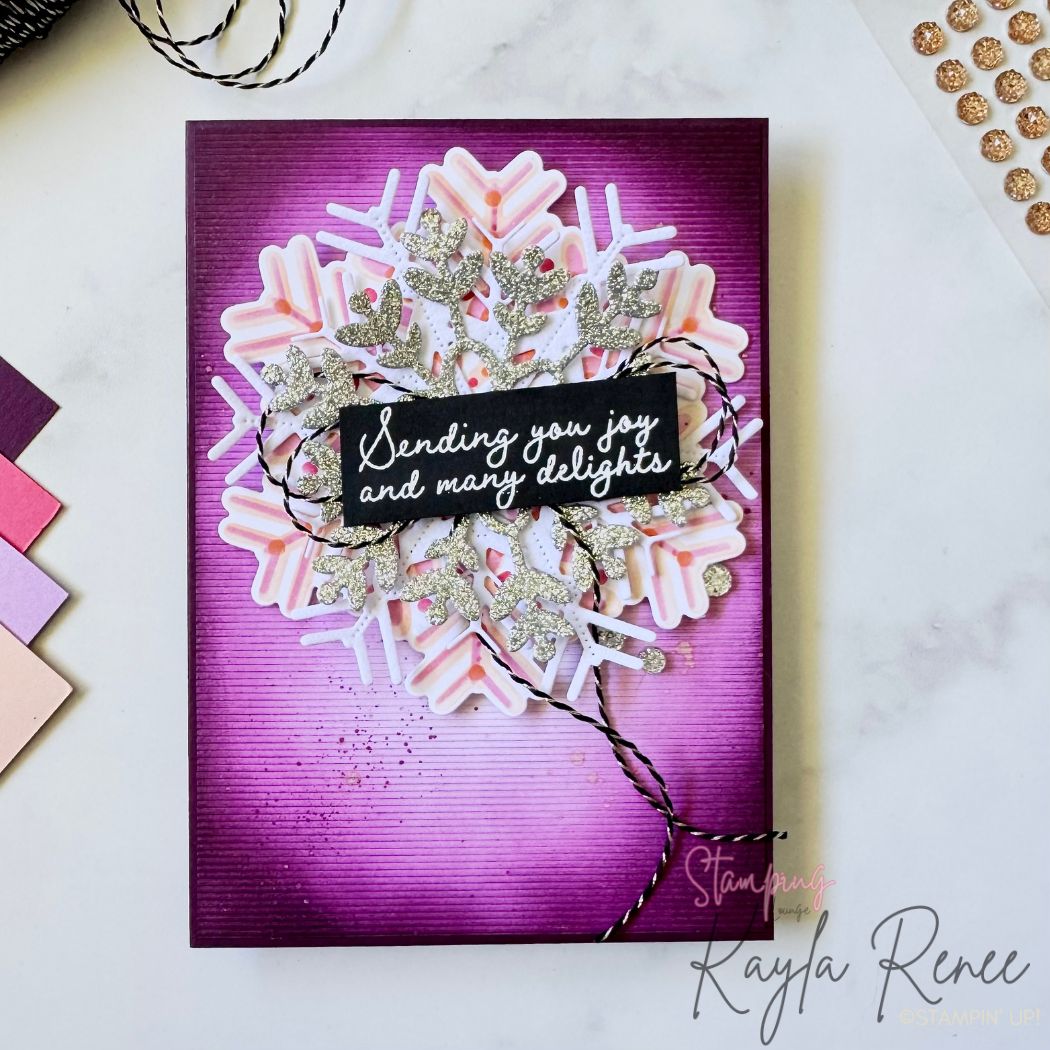 Flat-lay view of the completed card on a white surface. The deep-purple ink-blended background highlights a large layered snowflake with glitter silver and pink-white elements. Black-and-white twine wraps the centre, holding a sentiment panel reading “Sending you joy and many delights.” Small glitter enamel dots and coordinating pink-purple cardstock swatches are placed on the left of the scene.