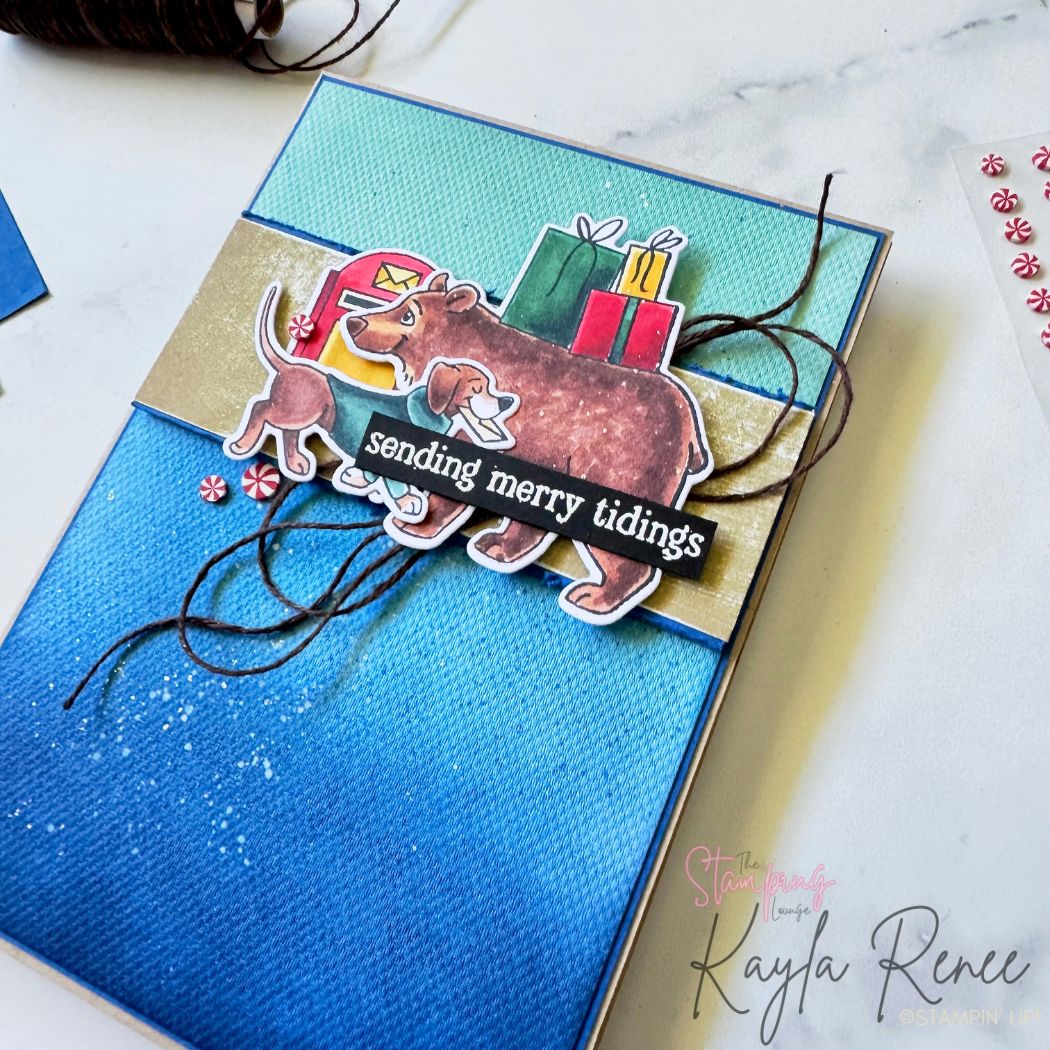 Close-up of a festive handmade card featuring a bear and puppy carrying envelopes, layered over presents and a red mailbox. The background is blue and gold with ink splatters and brown twine accents. Sentiment: 'sending merry tidings.
