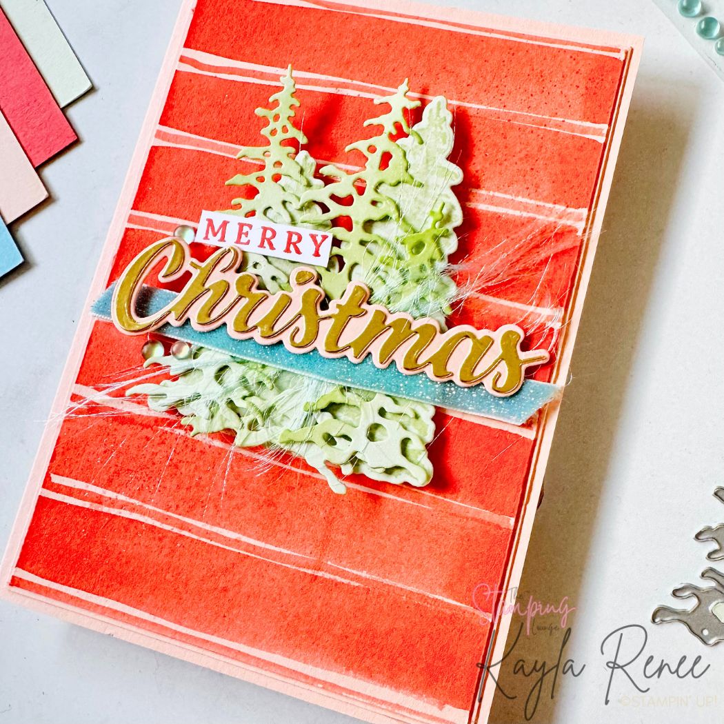 Close-up of the Christmas card showing layered pale green die-cut pine trees, gold-outlined “Christmas” sentiment on a blue banner, and fuzzy white thread behind the design. The red textured striped background is visible, with embellishments and dies in the scene.