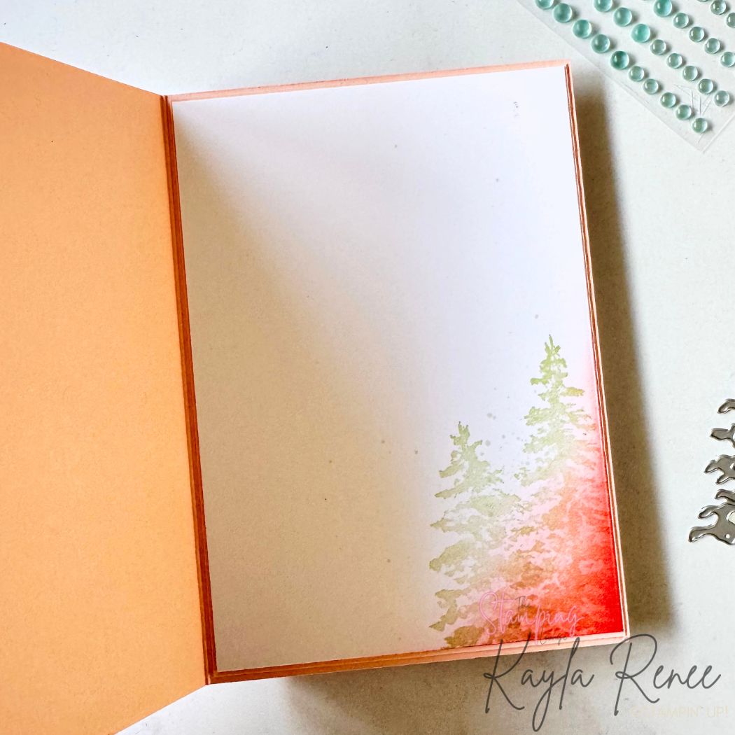 Inside of the card with a peach border and white insert panel. The lower right corner features softly ink-blended pine trees in green and red, creating a subtle winter forest scene.