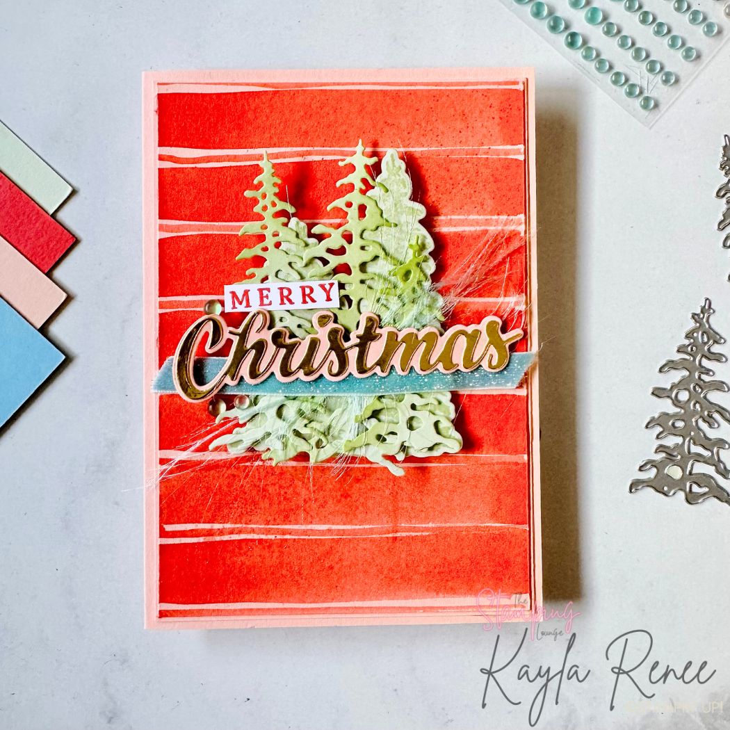 Handmade Christmas card featuring textured red watercolor stripes on a peach card base. Layered die-cut green pine trees sit behind a large gold-foiled “Christmas” sentiment with a small “Merry” label above it. Soft white fibers and subtle pearls add texture. Colour swatches and dies are visible beside the card.