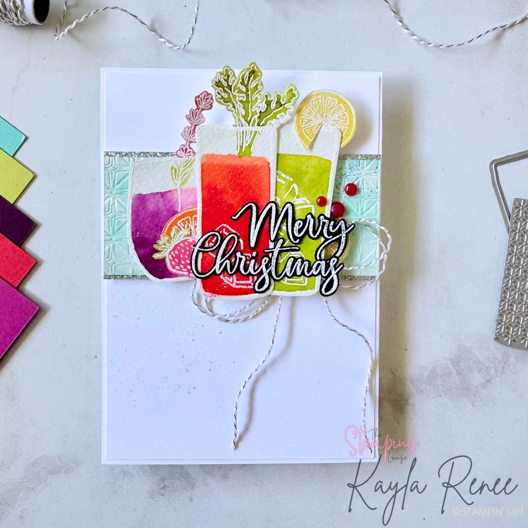 A handmade Christmas card featuring three colorful watercolor drinks in red, lime, and purple shades with embossed textured panels behind them. The drinks are decorated with citrus slices, berries, and leafy sprigs. The bold sentiment “Merry Christmas” is centered in black and white script, layered on twine. The background is crisp white with subtle ink splatter and playful festive embellishments.