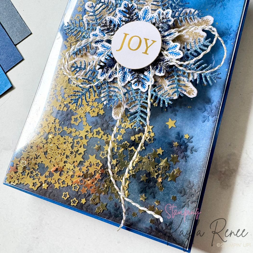 A zoomed-in view of the blue and gold Christmas shaker card showing the dimensional snowflake embellishment with glittery branches and navy stamped leaves. Gold star sequins fill the shaker pocket, and a white circle sentiment saying “JOY” sits on top. Textured inking adds depth to the winter scene.