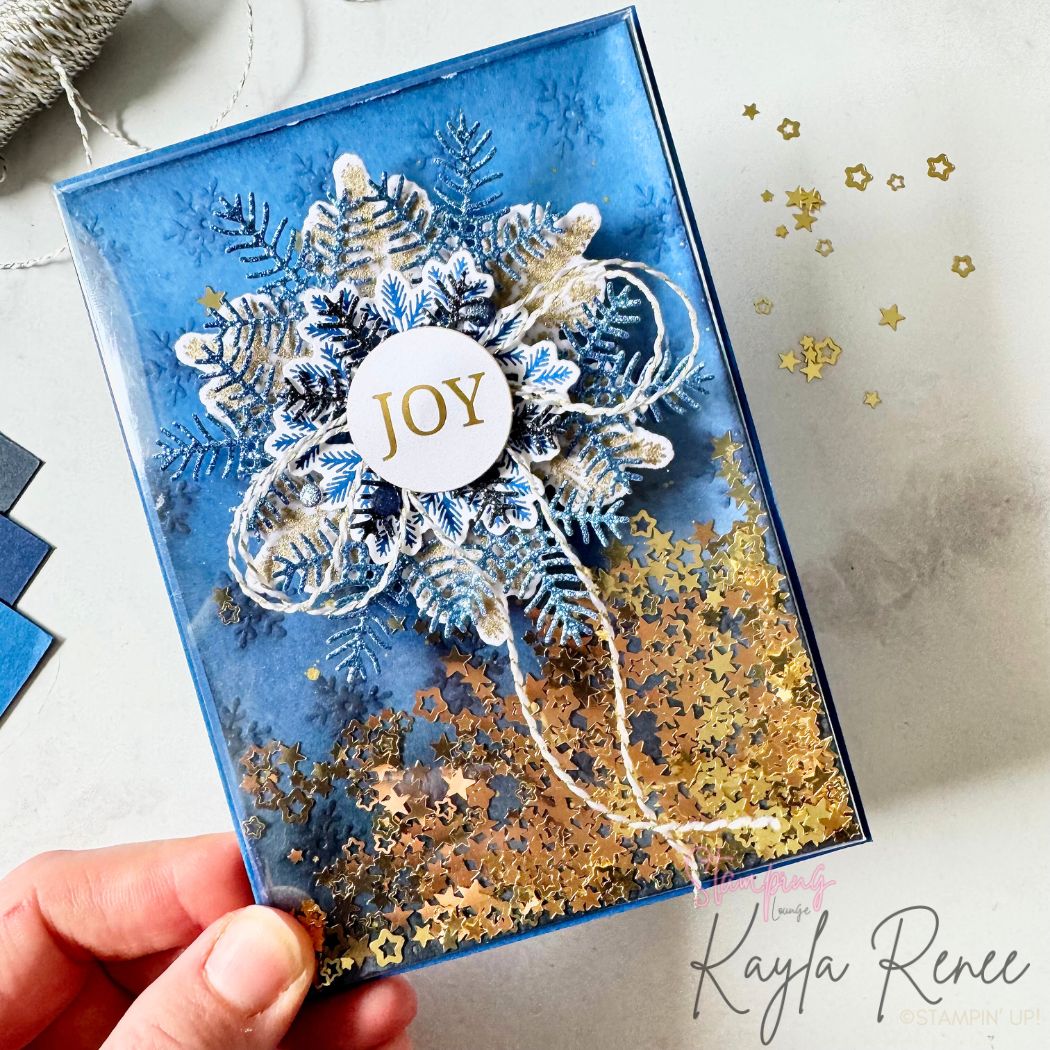 A hand holding the blue snowflake shaker card at an angle, showing the movement of gold star sequins. The snowflake layers, twine bow, and embossed icy blue background are visible, giving the card an elegant winter sparkle.