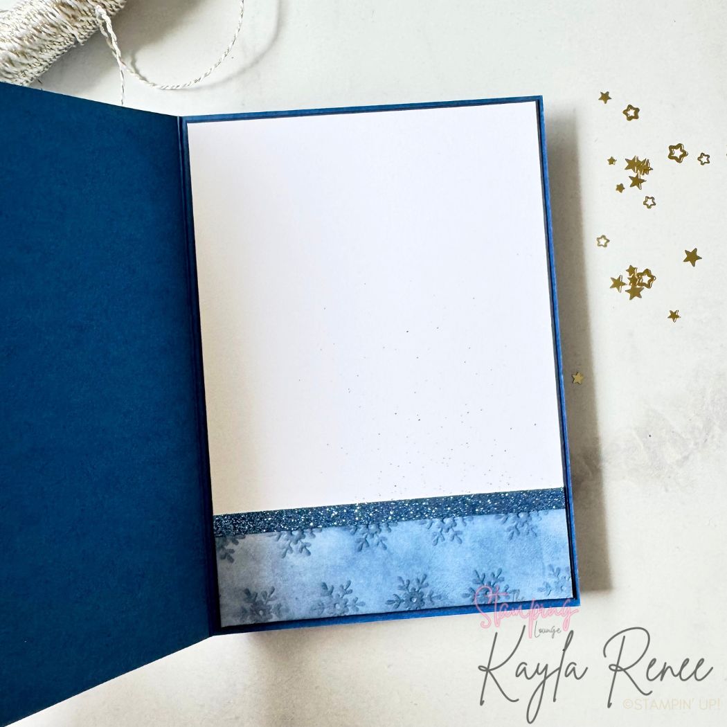 Inside of the handmade Christmas card with a deep blue border. The writing panel is white with subtle ink splatters, and a strip of blue snowflake patterned paper and glitter cardstock decorates the bottom edge for a cohesive winter design.