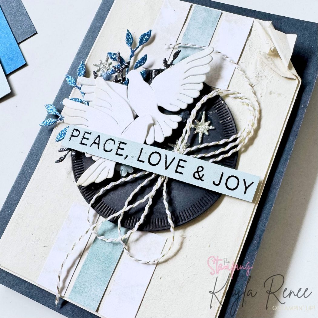 Close-up of the handmade dove Christmas card showing detailed die-cut doves, blue glitter leaves, textured black circle, and layered sentiment that reads “PEACE, LOVE & JOY.” Twine loops and small star embellishments add dimension over soft neutral and blue striped layers.