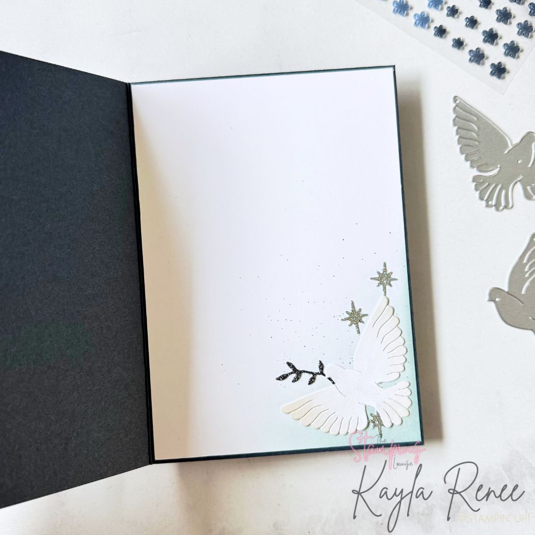 Inside of handmade Christmas card with clean white panel. Bottom corner features a soft blue ink-blended area with a white die-cut dove holding a branch and small glitter stars, with subtle ink splatter for texture.
