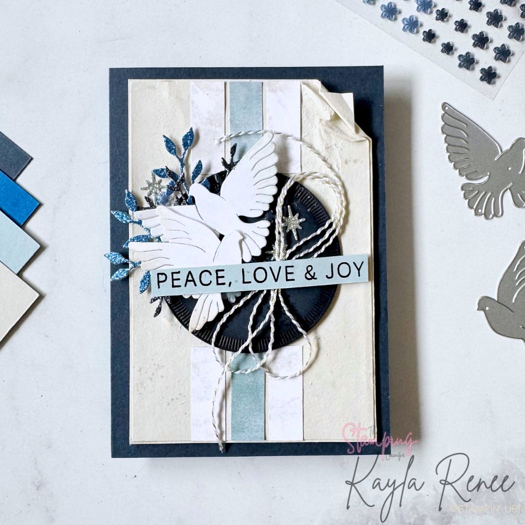 Handmade Christmas card in soft winter blues and creams featuring two layered white die-cut doves above a black textured circle wrapped with white twine. Blue glitter foliage and silver stars accent the scene, with a sentiment strip reading “PEACE, LOVE & JOY.” The background has distressed soft stripes and layered torn-edge paper for a vintage winter feel.