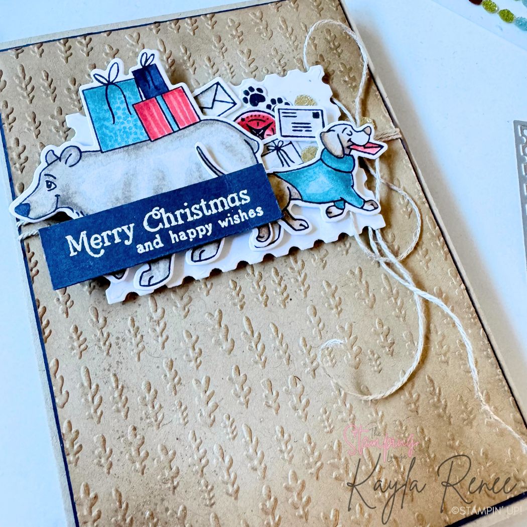 Close-up of handmade Christmas card featuring stamped and coloured polar bear and dachshund with holiday presents, layered over kraft embossed background and scalloped die cut. Navy heat-embossed sentiment reads “Merry Christmas and happy wishes,” accented with rustic linen thread twine.