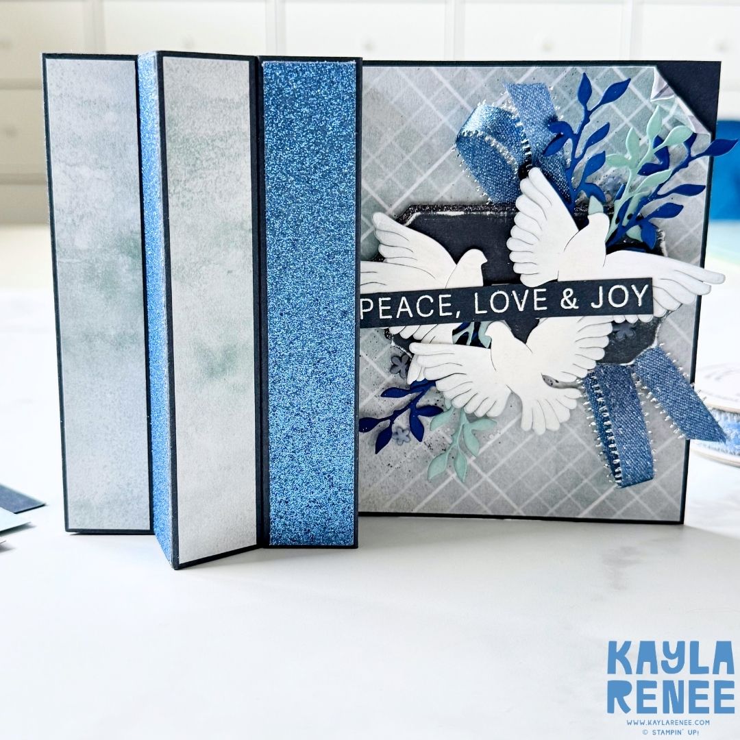 Front panel of “Peace, Love & Joy” handmade card by Kayla Renee from the Stampin’ Up! Peaceful Gardens suite. Designed with white doves, layered foliage, blue sparkle ribbon, and silver accents for an elegant holiday look.
