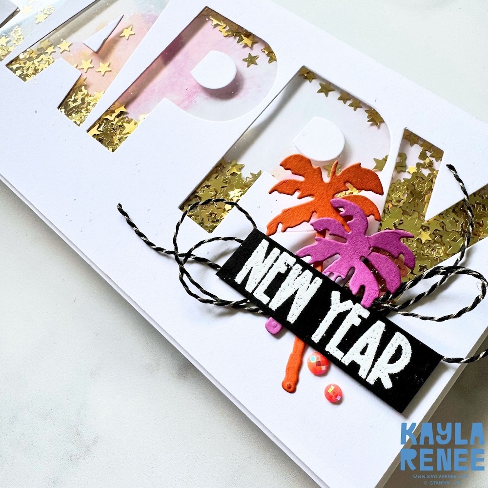 Close-up of a New Year shaker card showing gold star sequins behind the letter cut-outs, layered pink and gold palm tree die cuts, black-and-white twine, and a bold “NEW YEAR” sentiment strip.