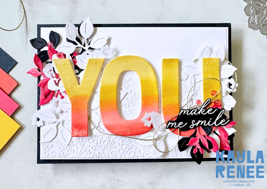 Handmade greeting card featuring large “YOU” letters created with Stampin’ Up! Party Alphabet dies, blended in a warm yellow-to-coral ombré. White layered florals and leaves frame the letters, with a black sentiment strip reading “make me smile” from the Charming Day bundle.