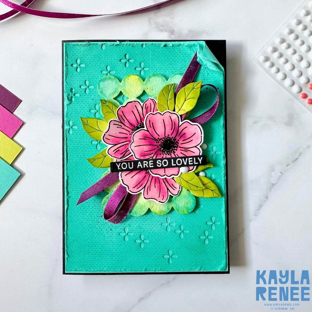 Teal handmade card featuring pink layered flowers, green leaves and a “You Are So Lovely” sentiment with embossed background and ribbon accent.