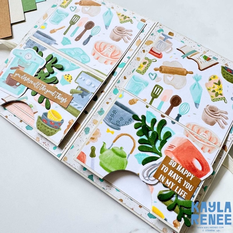 Top view of interactive papercraft card with pull-down sections opened, showing decorative kitchen imagery, pastel patterns, and a blank journaling panel with a small teal heart.