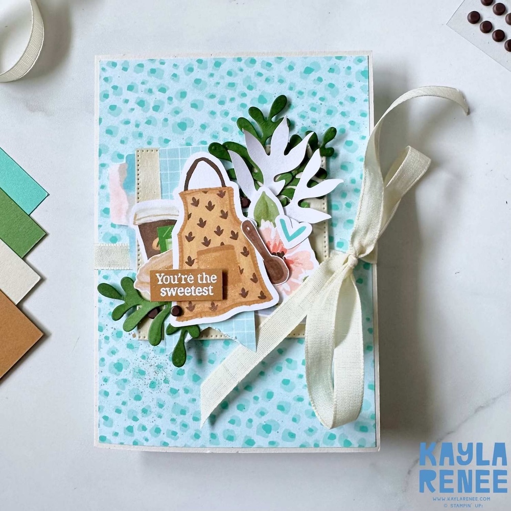 Handmade cooking-themed card featuring a teal speckled background, apron and coffee cup die cuts, leafy greenery, and a cream ribbon tied along the side with the sentiment “You’re the sweetest.