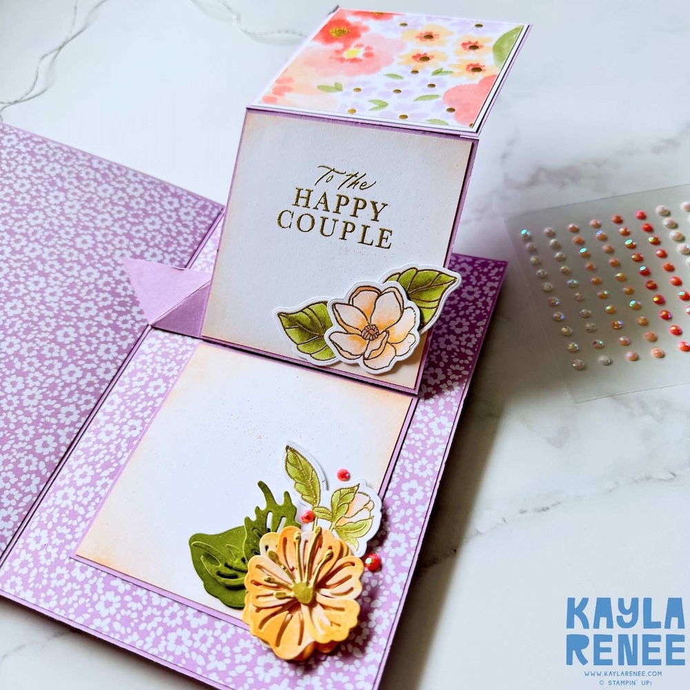 Close-up of expanding pop-up wedding card featuring a stamped sentiment “To the Happy Couple” with peach floral embellishments and pastel patterned paper.