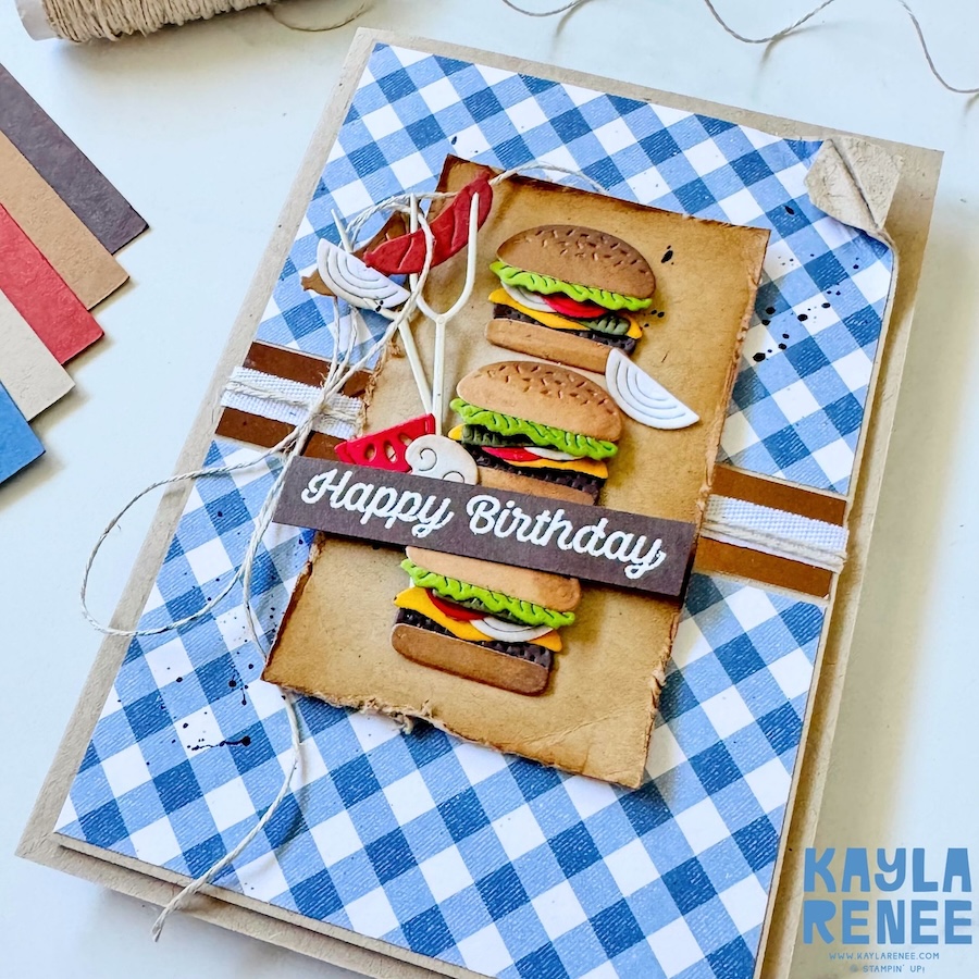 Angled view of handmade BBQ-themed birthday card with distressed cardstock layers, burger die cuts, and cooking utensil embellishments over blue gingham patterned paper.