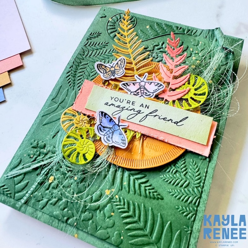 Angled detail view of the card highlighting dimensional layers, including die-cut ferns, circular elements, butterflies, and soft fibres over a textured green embossed background.