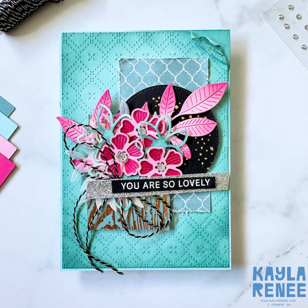 Full front view of a handmade card created using The Stamping Lounge Sketch Box, showcasing a layered floral arrangement with pink die-cut flowers, soft aqua leaves, patterned paper, and a black circle with gold splatter accents on an embossed aqua card base.