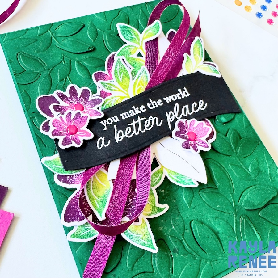 Angled view of a Stampin’ Up! Garden Poetry Suite handmade card showing dimensional pink and purple flowers, leafy accents, and a purple ribbon layered beneath a bold black sentiment strip.