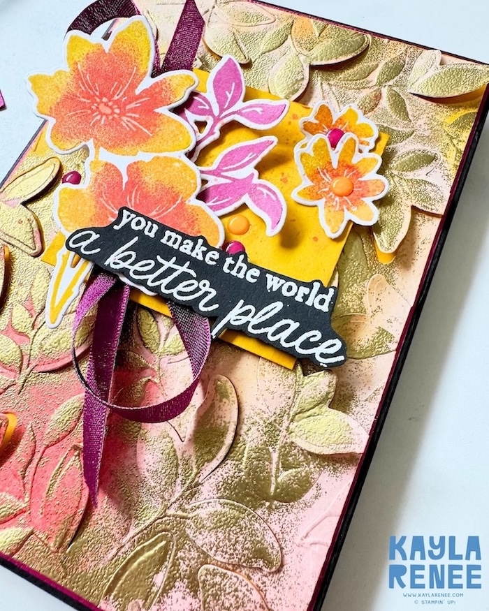 Close-up of a Stampin’ Up! Garden Poetry Suite card created for GDP550, highlighting the dimensional dry embossed and heat embossed leafy background. Multi-colour stamped flowers in vibrant yellow, orange, and coral tones are layered with bright pink foliage, ribbon loops, and enamel embellishments around the sentiment, “You make the world a better place.”